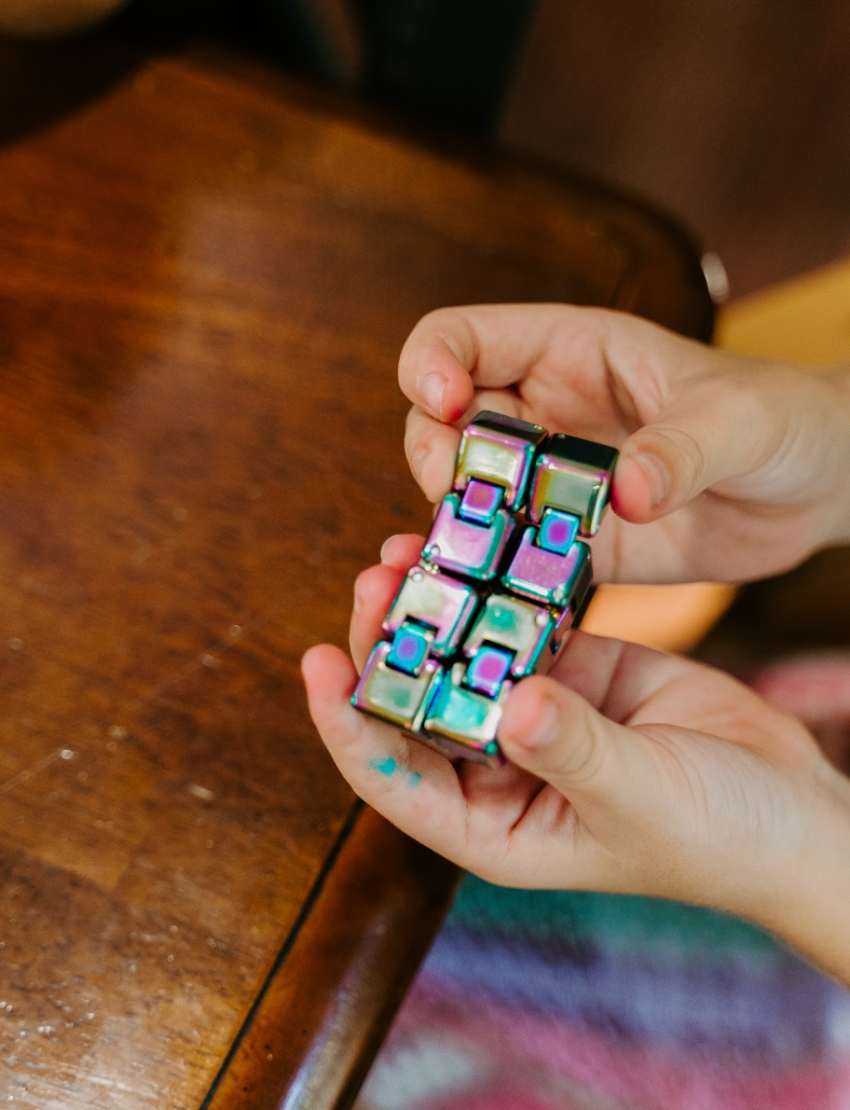 The "Understanding My Regulation Preferences Kit" by The Play Way is a green and teal box filled with various sensory and stress relief products, including a pack of resistance bands, an infinity cube, a box of Aaron's Thinking Putty, and a Hide Inside toy. Additionally, it comes with a regulation tracker sheet.