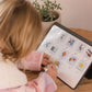 A parent with two kids is on a bed. They and one child are exploring The Play Way's Sleep Program box, while the other child holds a book. The bedroom features a decorative headboard and a patterned lamp.