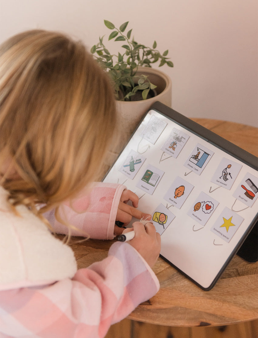 A parent with two kids is on a bed. They and one child are exploring The Play Way's Sleep Program box, while the other child holds a book. The bedroom features a decorative headboard and a patterned lamp.