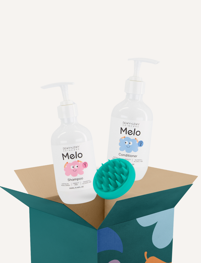 Dewy + Lewy Melo shampoo, conditioner, and scalp massager emerging from an open gift box.