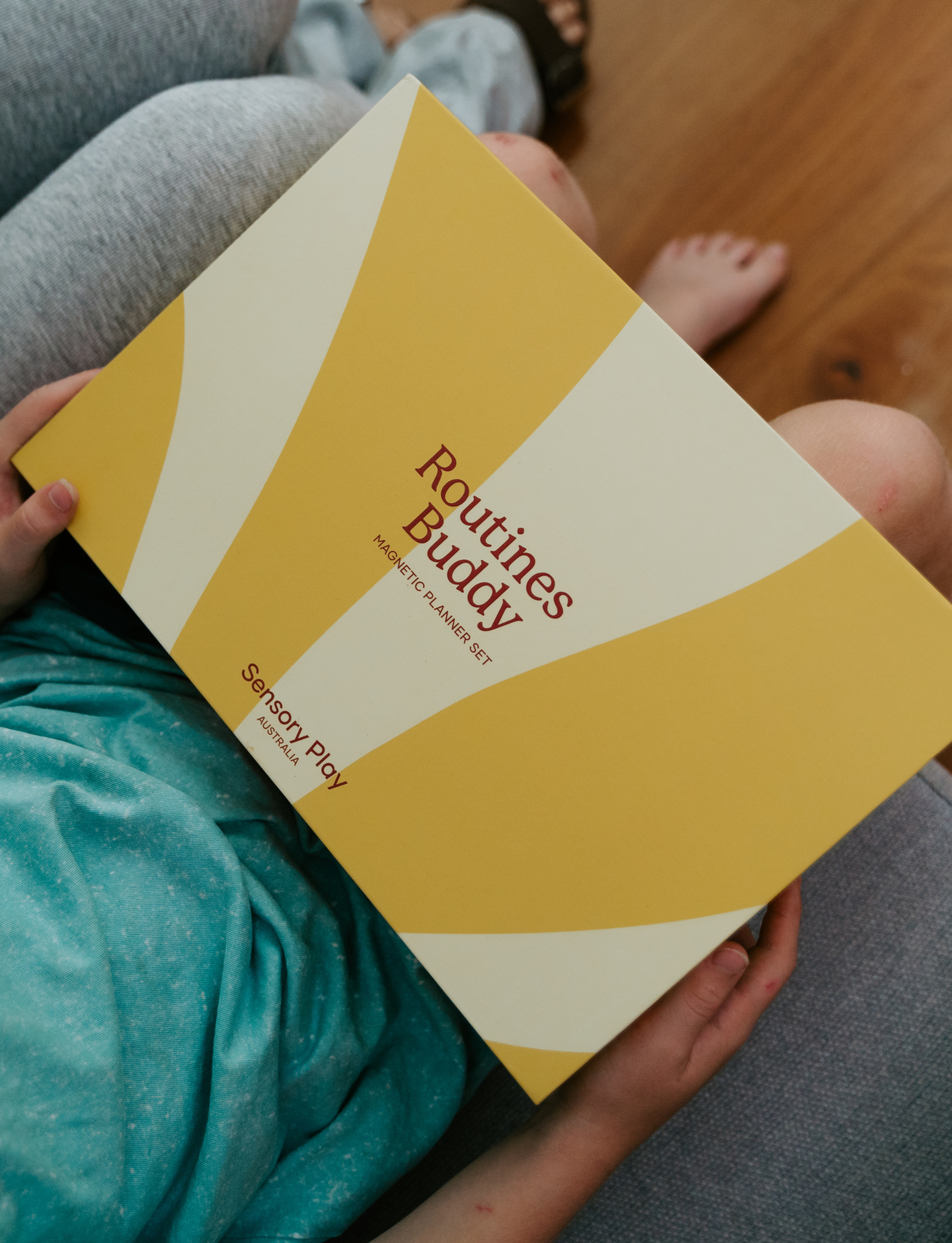 Lifestyle image of a child holding the yellow box of the Routines Buddy Magnetic Planner Set on their lap. A toolkit from Sensory Play Australia designed to empower children and families with daily routines.
