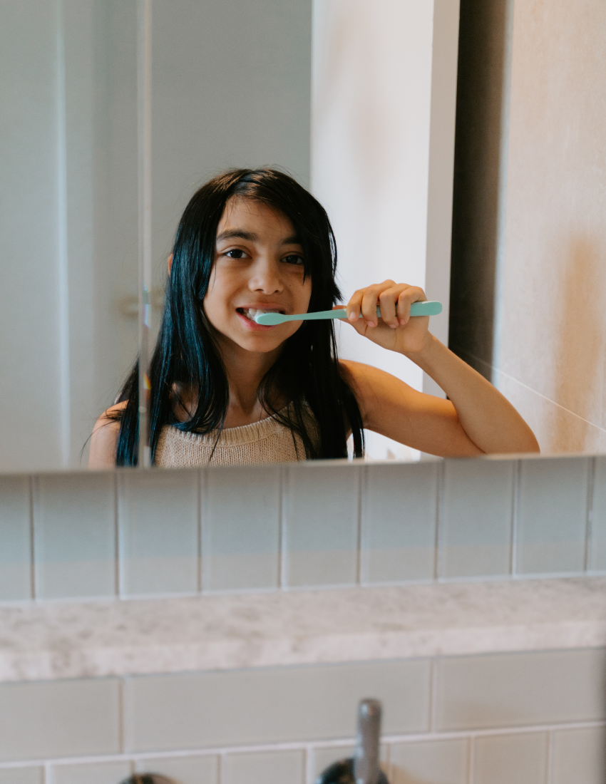 A person with long dark hair is brushing their teeth in front of a bathroom mirror, surrounded by white tiles and a light-colored countertop. They are holding a blue-handled toothbrush and wearing a beige top. This scene reflects the themes taught in The Play Way's Sensory: Self-Care and Considerations Masterclass.