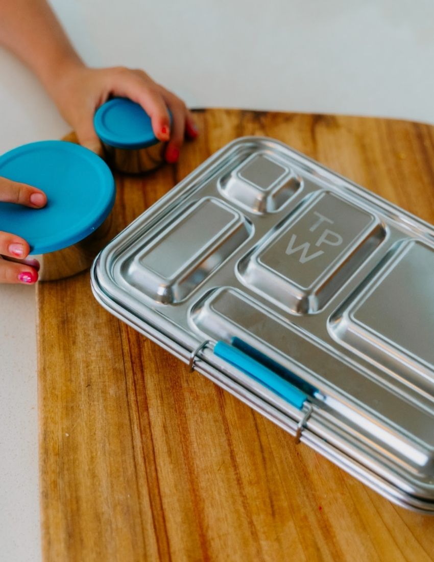 Introducing the Lunch Buddy Bento Set by The Play Way, a sleek rectangular stainless steel lunchbox with multiple compartments. The top right section is larger than the others, with "TPW" engraved in the central compartment. It features a silicone seal lid, rounded edge design, clasp closure, and a polished metallic finish.