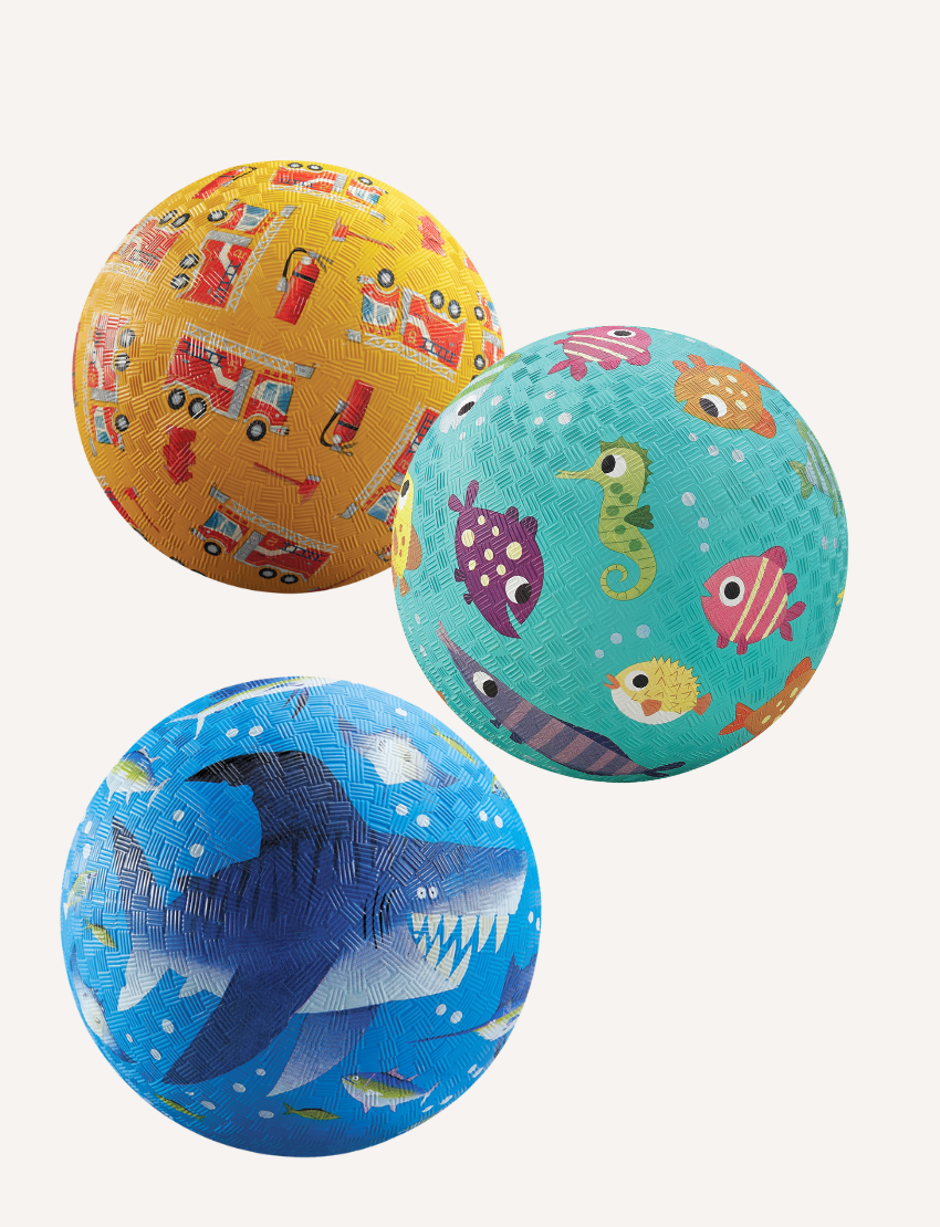 Three Crocodile Creek 7 Inch Playground Balls are displayed: a yellow one with fire trucks, a teal ball with sea creatures like fish and seahorses, and a blue one with shark designs—ideal for boosting children's gross motor skills.