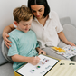 A mother and son sit together on a couch using the Routines Buddy visual schedule board. The child writes on the board, showing how the tool fosters connection and interactive, play-based learning for daily routines.