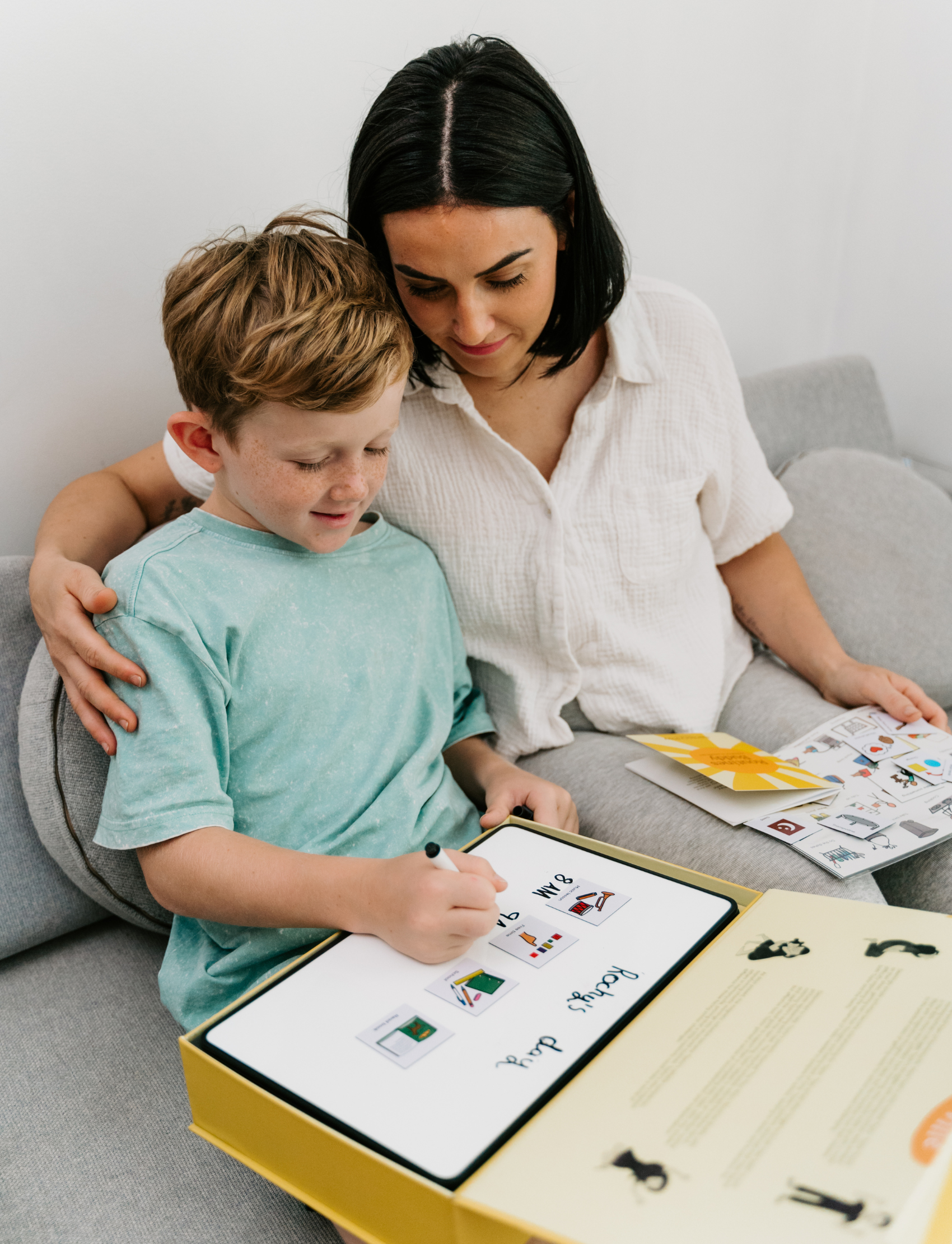 A mother and son sit together on a couch using the Routines Buddy visual schedule board. The child writes on the board, showing how the tool fosters connection and interactive, play-based learning for daily routines.