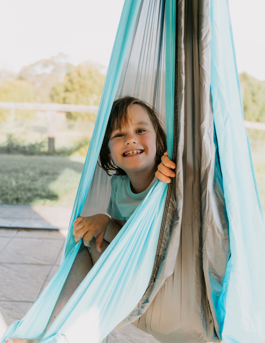 A child with short brown hair smiles while sitting in a bright blue hammock outdoors. The light fabric of the hammock contrasts nicely with the grassy area and trees in the background. The child is wearing a light-colored shirt, enjoying their time as they experience The Play Way's Sensory: An Overview Masterclass-inspired play session.