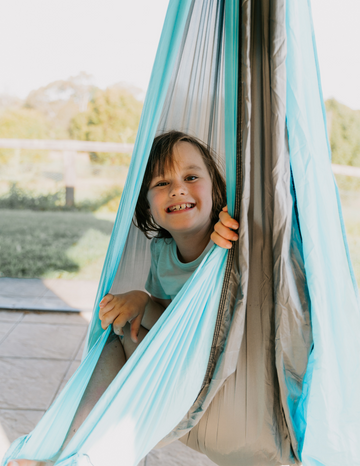 A child with short brown hair smiles while sitting in a bright blue hammock outdoors. The light fabric of the hammock contrasts nicely with the grassy area and trees in the background. The child is wearing a light-colored shirt, enjoying their time as they experience The Play Way's Sensory: An Overview Masterclass-inspired play session.