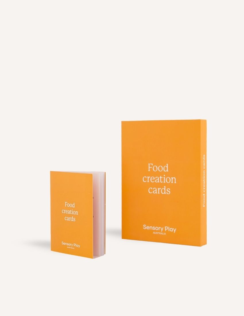 The image features two items: a smaller, open book and a larger, closed box, both with plain yellow-orange covers and white text that says "Food Creation Card Set." These items are branded with "Sensory Play Australia" at the bottom. The Food Creation Card Set in Box is designed to enhance your mealtime experience.