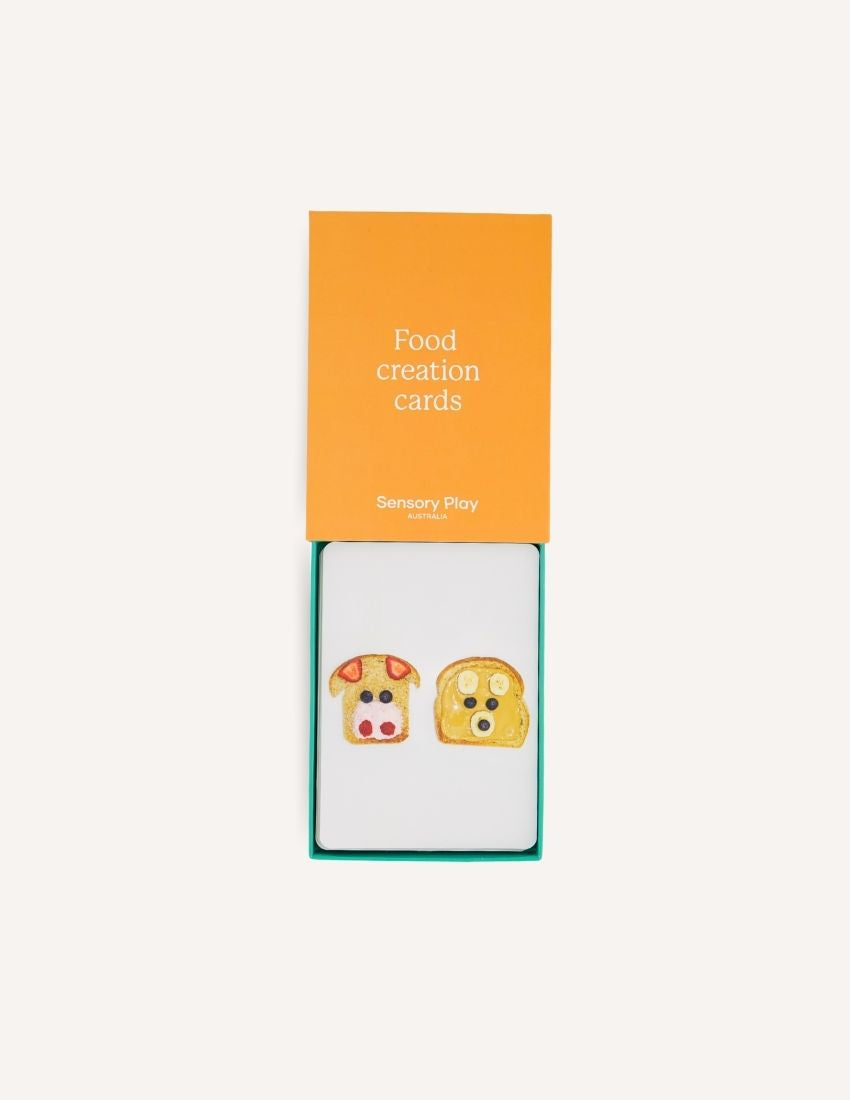 The image features two items: a smaller, open book and a larger, closed box, both with plain yellow-orange covers and white text that says "Food Creation Card Set." These items are branded with "Sensory Play Australia" at the bottom. The Food Creation Card Set in Box is designed to enhance your mealtime experience.