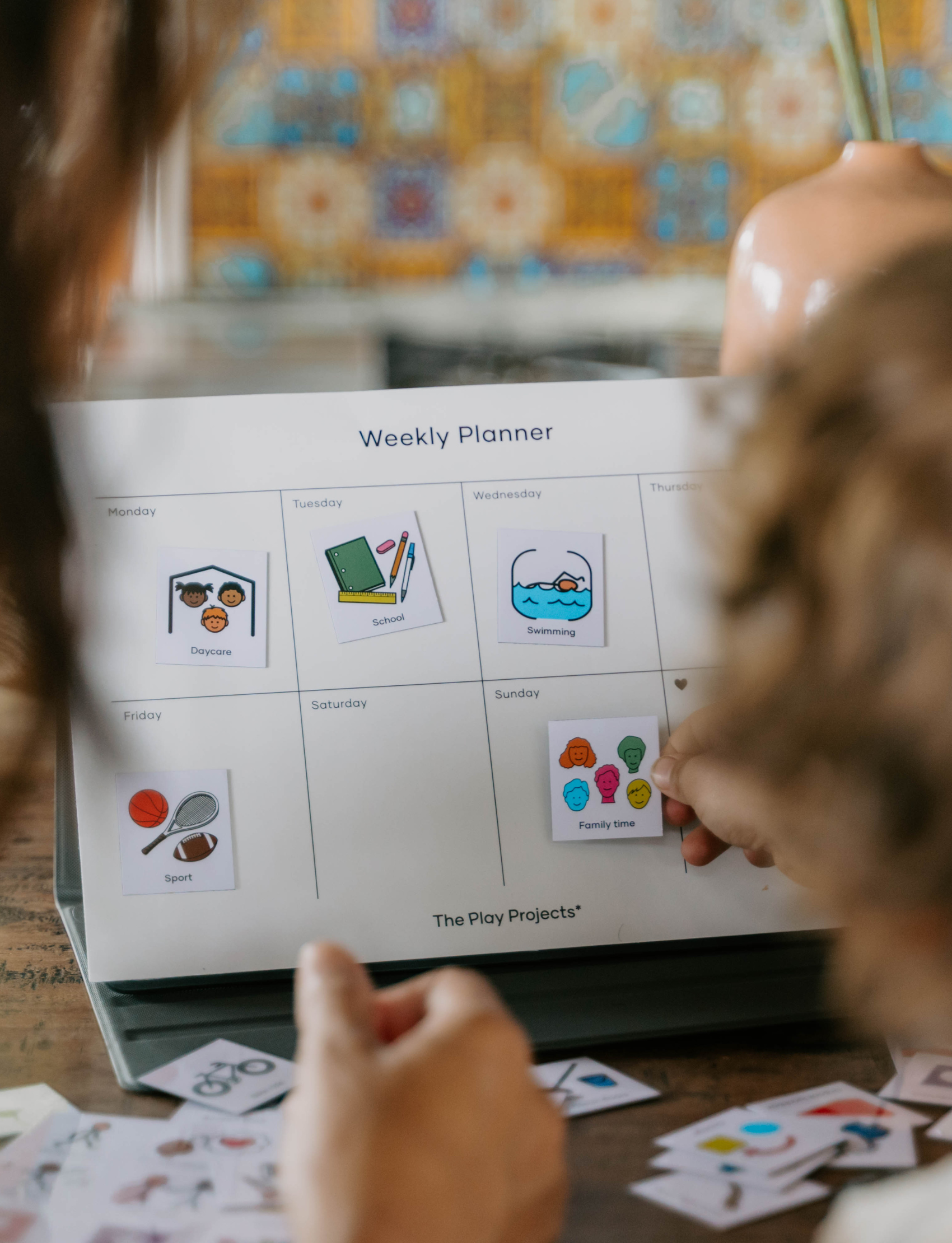 A child's hand places a "Family time" picture icon onto a magnetic Weekly Planner. This visual aid helps children understand their schedule with icons for school and sports, reducing anxiety by building a clear routine.