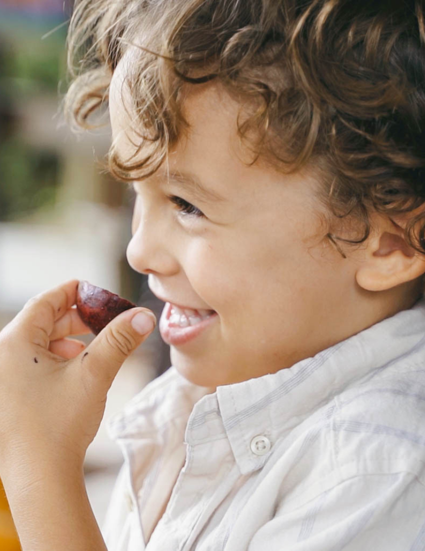 Close-up of a smiling child being fed a piece of fruit by a hand.