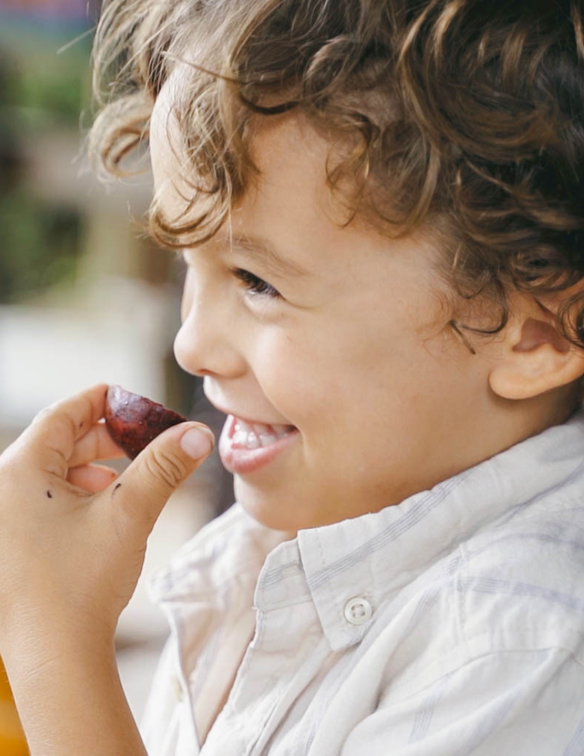 A young child with curly hair smiles while holding a small piece of fruit close to their mouth, embodying the joy promoted by The Play Way's Feeding Masterclass. They are wearing a light-colored shirt, and the background is artistically blurred with neutral and green tones.