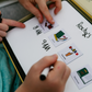 Close-up of a child and parent using the Routines Buddy whiteboard. They arrange picture icons next to handwritten times, creating a personalised daily schedule to teach time management and foster independence.