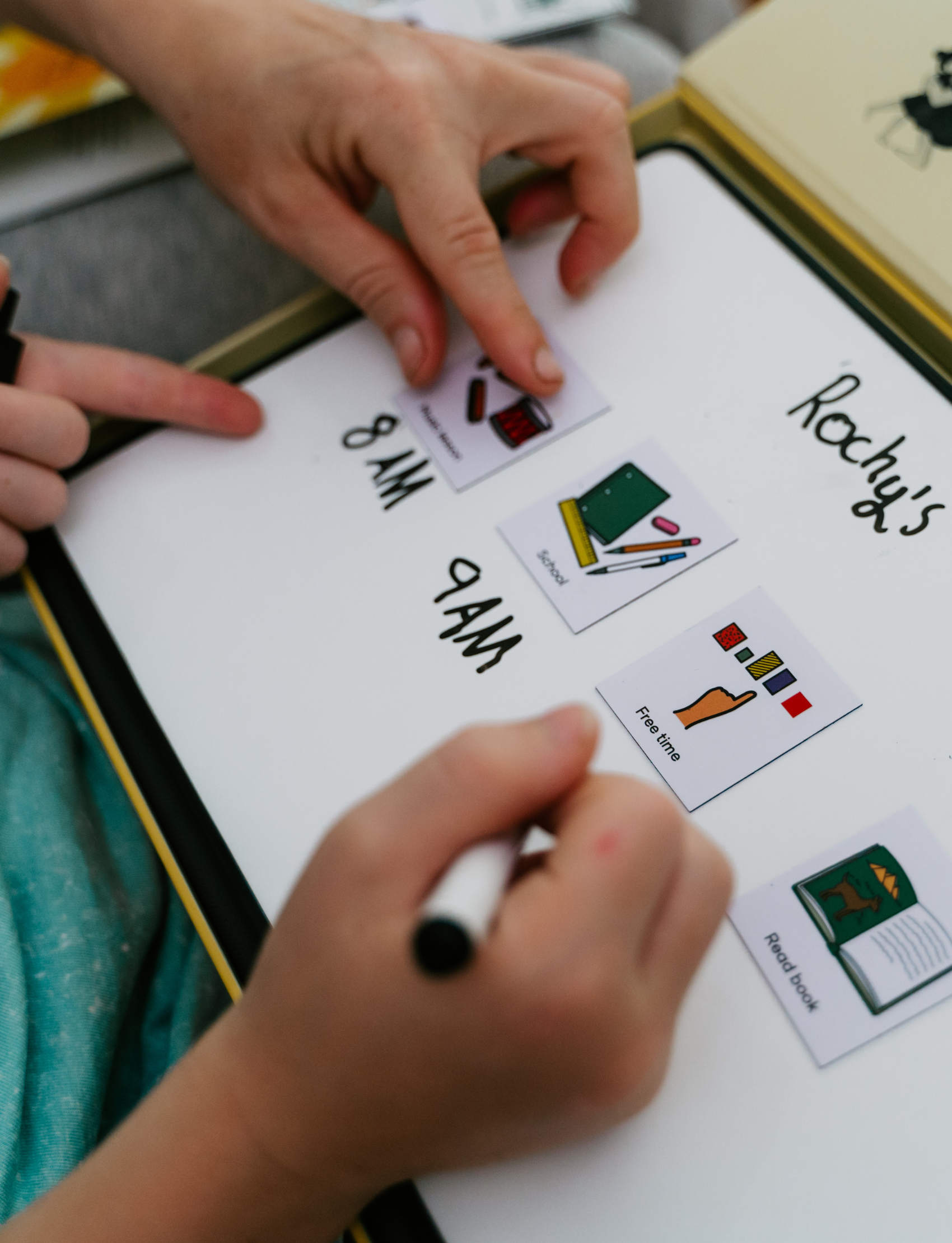 Close-up of a child and parent using the Routines Buddy whiteboard. They arrange picture icons next to handwritten times, creating a personalised daily schedule to teach time management and foster independence.