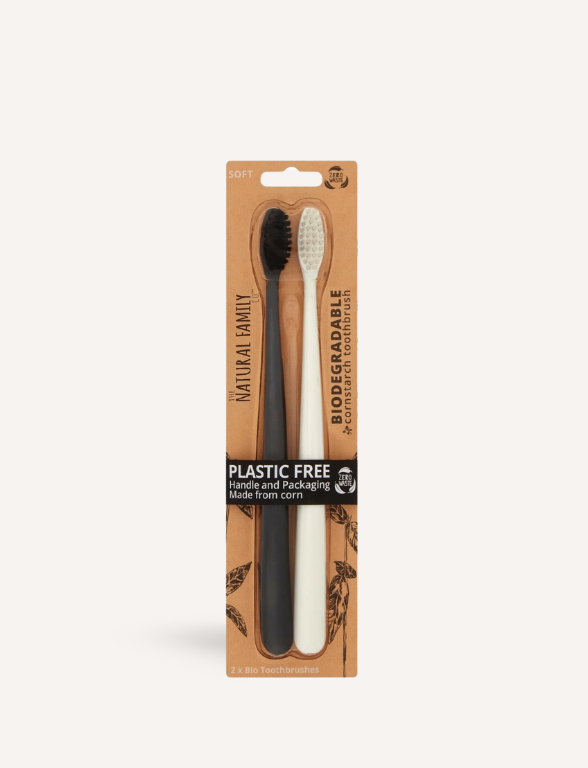 The Bio Toothbrush - Mixed Twin Pack by Jack N' Jill includes two biodegradable toothbrushes, one black and one white with soft bristles. They're plastic-free and made from corn, packaged with a backdrop of decorative branches with white buds.