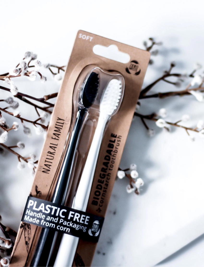The Bio Toothbrush - Mixed Twin Pack by Jack N' Jill includes two biodegradable toothbrushes, one black and one white with soft bristles. They're plastic-free and made from corn, packaged with a backdrop of decorative branches with white buds.