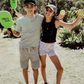 Two children sit on grass by an open cardboard box from The Play Way's "Building Motor Skills through Play Program," featuring various items. Holding green paddles with text, they wear hats and casual clothes. Bushes and trees in the background suggest a park setting.
