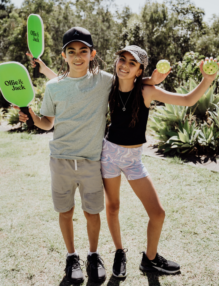 Two children sit on grass by an open cardboard box from The Play Way's "Building Motor Skills through Play Program," featuring various items. Holding green paddles with text, they wear hats and casual clothes. Bushes and trees in the background suggest a park setting.