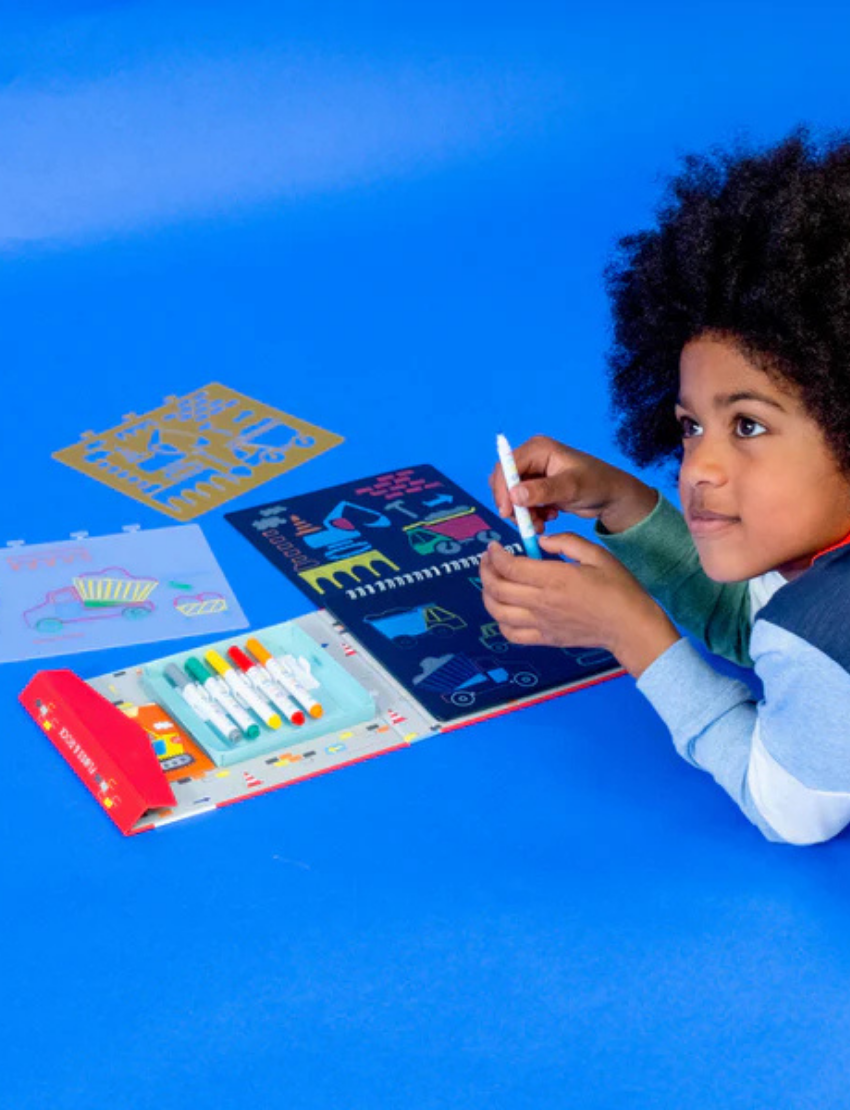 A child with curly hair lies on their stomach on a blue background, focused intently. In front of them are a Floss & Rock Chalk Board Sketchbook, markers, and various stencils. The child holds a marker, fully engrossed in the creative activity.