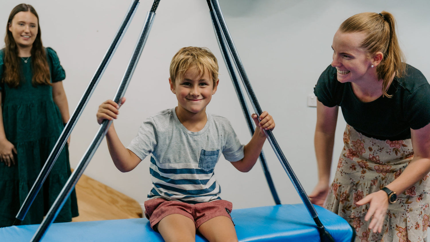 Child sitting on a blue platform with support, supervised by two adults in a gym setting.