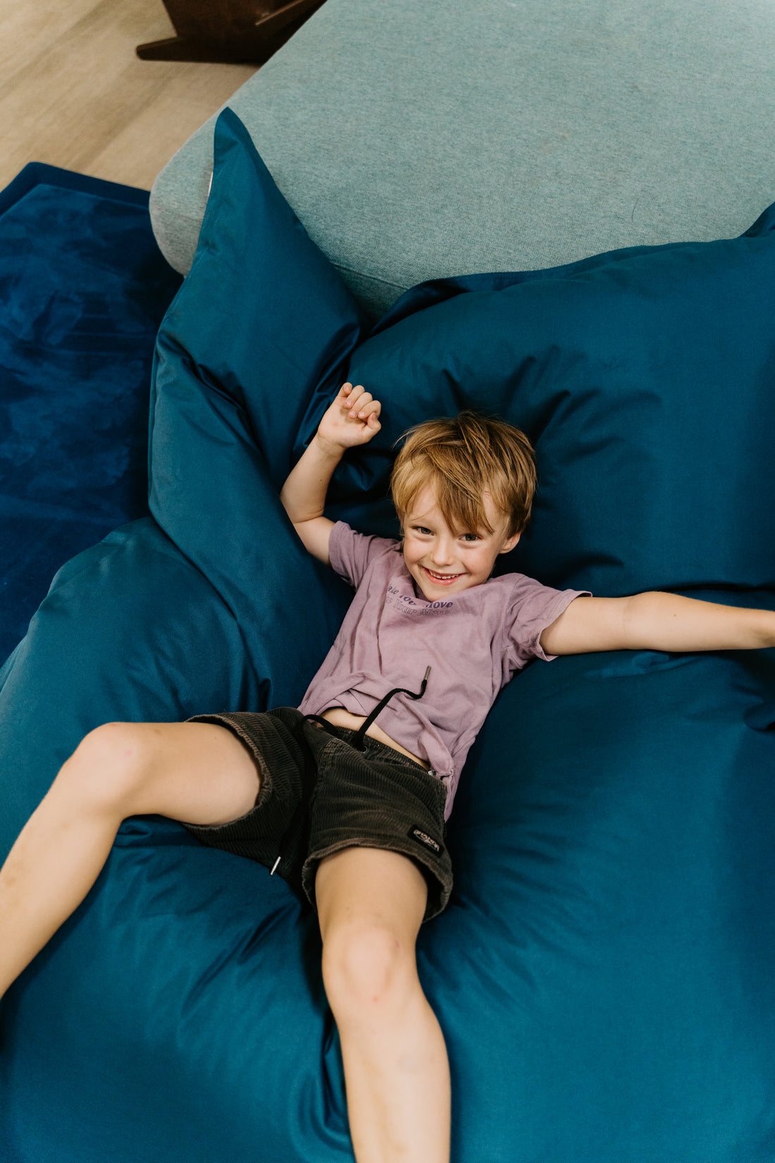 Two children immerse themselves in imaginative play on a large, blue Crash Mat by Sensory Play Australia, positioned in front of a gray sofa. Over the sofa's backrest hang colorful shirts, with one child sporting a vibrant tie-dye shirt and the other clad in a light blue shirt and shorts. Their activities encourage the development of motor skills through fun and creativity.