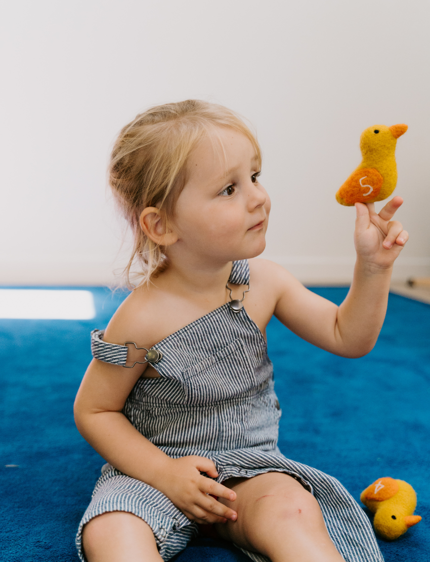 A child, wearing a striped outfit with a shoulder strap off, sits on a blue carpet. They curiously gaze at their small yellow and orange stuffed bird toy from The Play Way's Early Years Language Program. Another similar toy rests nearby on the floor.