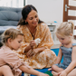 A child, wearing a striped outfit with a shoulder strap off, sits on a blue carpet. They curiously gaze at their small yellow and orange stuffed bird toy from The Play Way's Early Years Language Program. Another similar toy rests nearby on the floor.