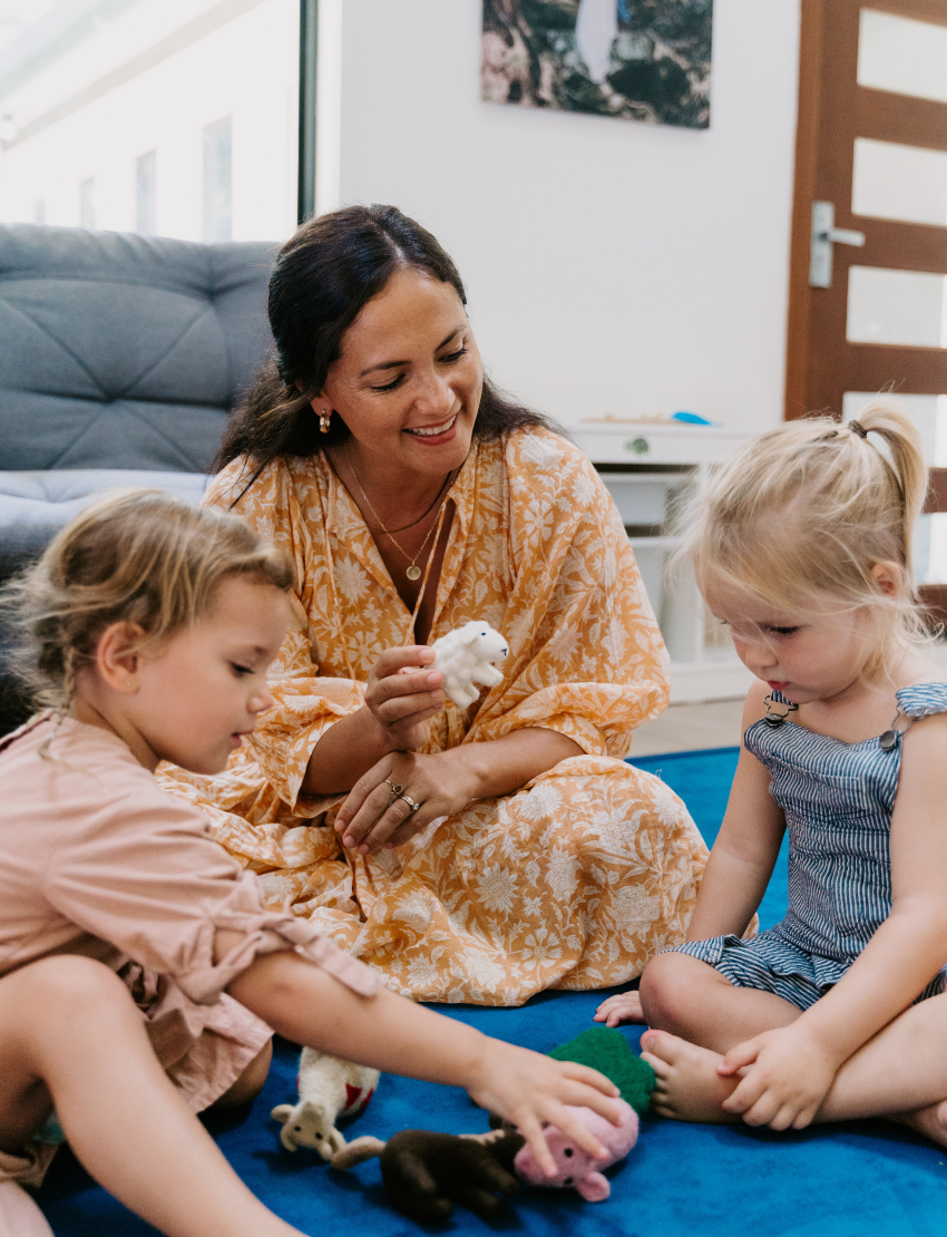 A child, wearing a striped outfit with a shoulder strap off, sits on a blue carpet. They curiously gaze at their small yellow and orange stuffed bird toy from The Play Way's Early Years Language Program. Another similar toy rests nearby on the floor.