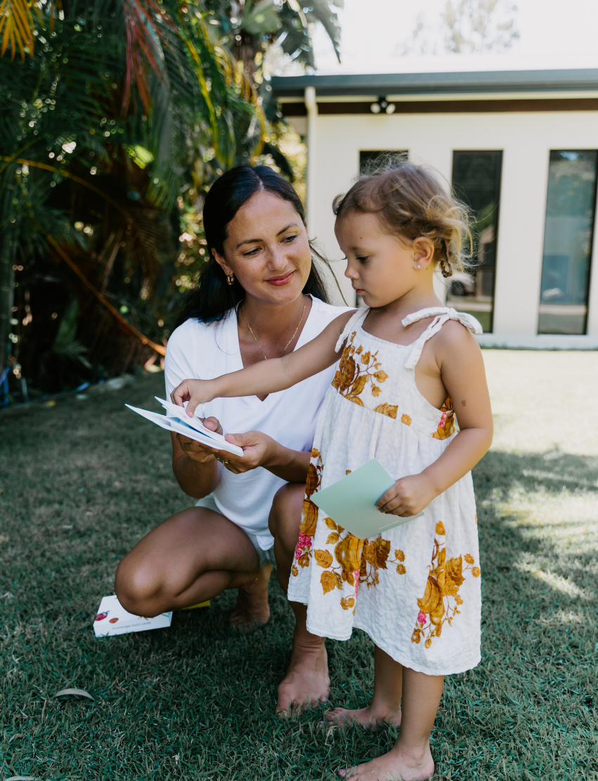 A child, wearing a striped outfit with a shoulder strap off, sits on a blue carpet. They curiously gaze at their small yellow and orange stuffed bird toy from The Play Way's Early Years Language Program. Another similar toy rests nearby on the floor.