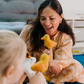 A child, wearing a striped outfit with a shoulder strap off, sits on a blue carpet. They curiously gaze at their small yellow and orange stuffed bird toy from The Play Way's Early Years Language Program. Another similar toy rests nearby on the floor.