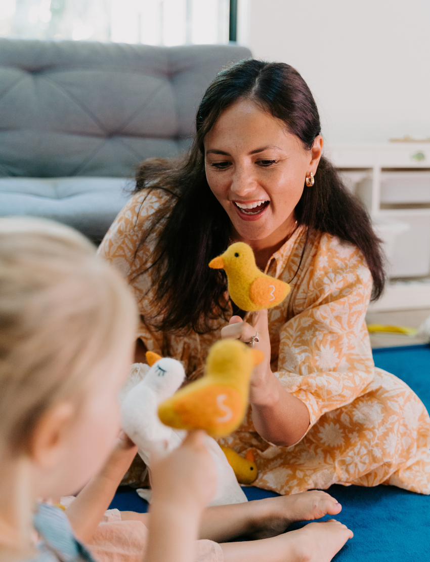 A child, wearing a striped outfit with a shoulder strap off, sits on a blue carpet. They curiously gaze at their small yellow and orange stuffed bird toy from The Play Way's Early Years Language Program. Another similar toy rests nearby on the floor.