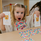 A young child with pigtails, looking surprised, holds a flashcard from The Play Way's "Emotions Part 1: Naming our Feelings Program," featuring a surprised face. She sits at a table with colorful polka-dot cards, and a background shelf displays toys and a globe.