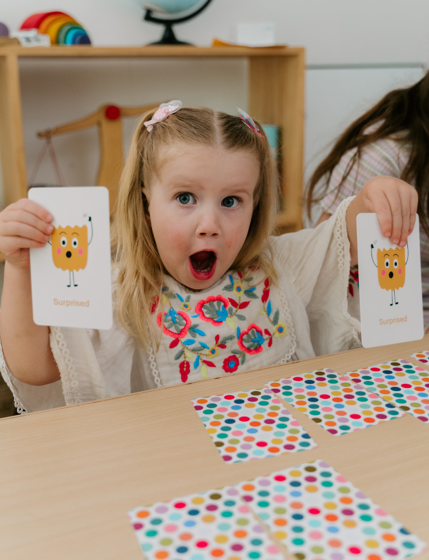 A young child with pigtails, looking surprised, holds a flashcard from The Play Way's "Emotions Part 1: Naming our Feelings Program," featuring a surprised face. She sits at a table with colorful polka-dot cards, and a background shelf displays toys and a globe.