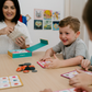 A woman and two children are at a table playing "Emotions Part 1: Naming our Feelings Program" by The Play Way. The woman holds a pouch, while a smiling child displays a card with pictures. The game box is open, and colorful images decorate the background wall.