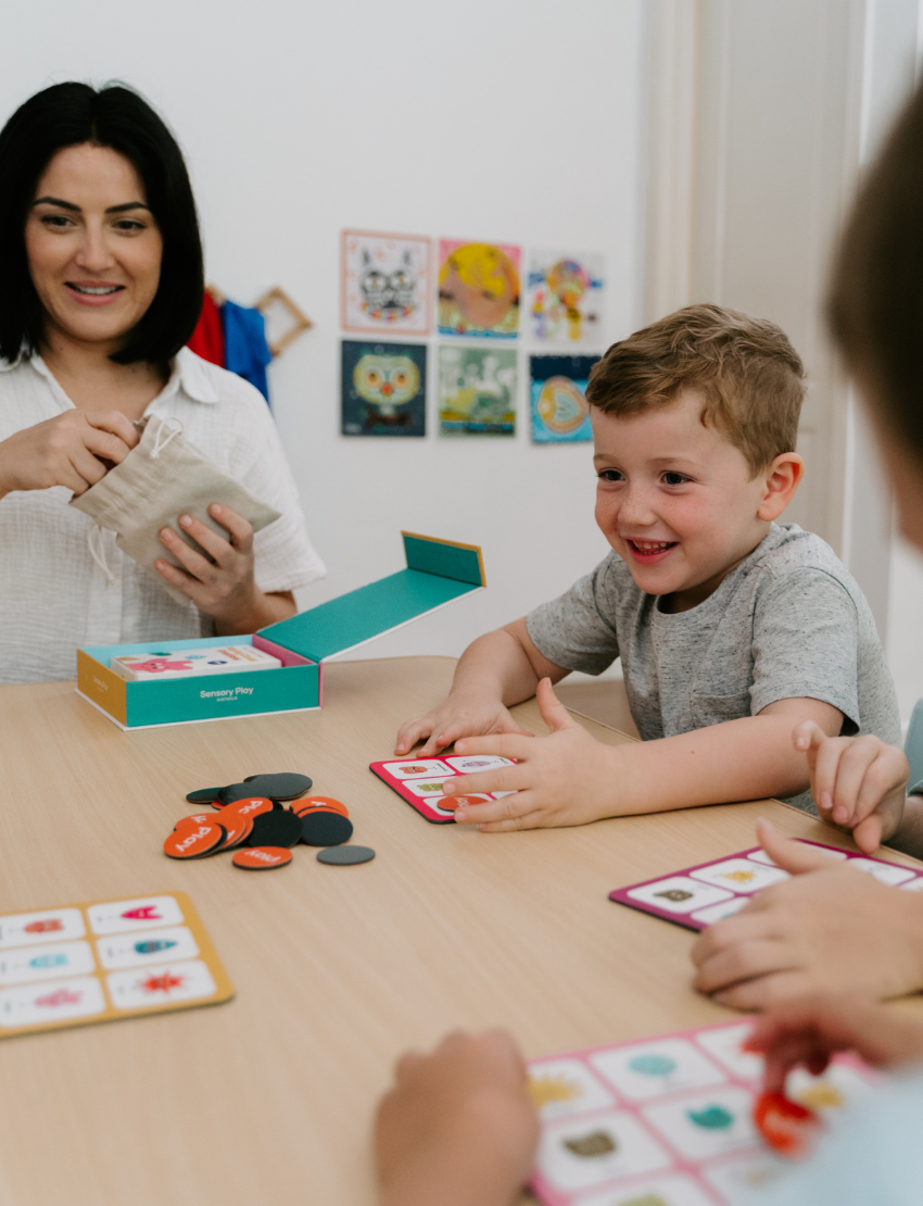 A woman and two children are at a table playing "Emotions Part 1: Naming our Feelings Program" by The Play Way. The woman holds a pouch, while a smiling child displays a card with pictures. The game box is open, and colorful images decorate the background wall.
