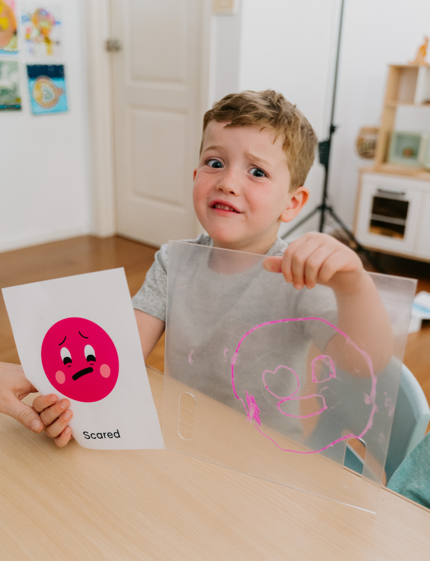 A woman and two children are at a table playing "Emotions Part 1: Naming our Feelings Program" by The Play Way. The woman holds a pouch, while a smiling child displays a card with pictures. The game box is open, and colorful images decorate the background wall.
