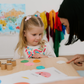 A woman and two children are at a table playing "Emotions Part 1: Naming our Feelings Program" by The Play Way. The woman holds a pouch, while a smiling child displays a card with pictures. The game box is open, and colorful images decorate the background wall.