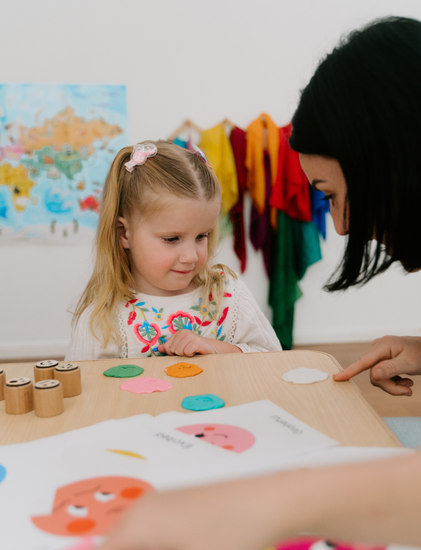 A woman and two children are at a table playing "Emotions Part 1: Naming our Feelings Program" by The Play Way. The woman holds a pouch, while a smiling child displays a card with pictures. The game box is open, and colorful images decorate the background wall.