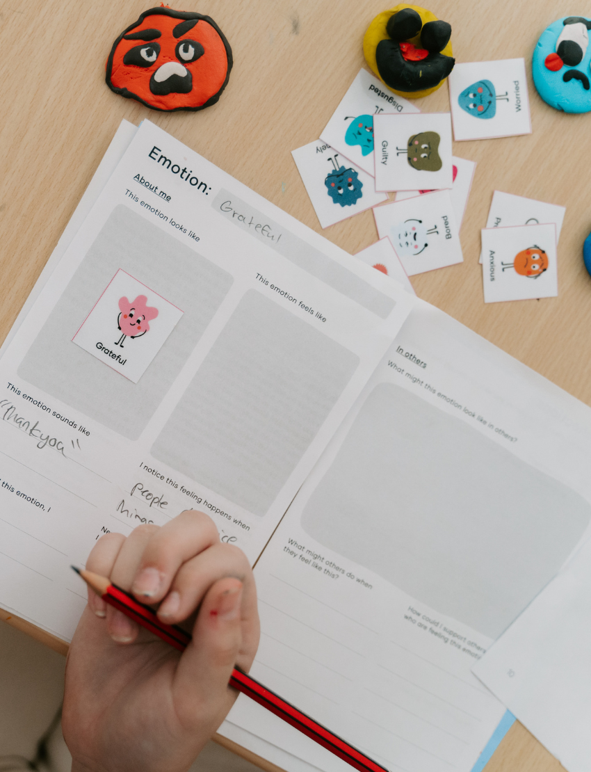 A woman and two children are at a table playing "Emotions Part 1: Naming our Feelings Program" by The Play Way. The woman holds a pouch, while a smiling child displays a card with pictures. The game box is open, and colorful images decorate the background wall.