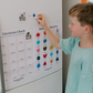 A woman and two children are at a table playing "Emotions Part 1: Naming our Feelings Program" by The Play Way. The woman holds a pouch, while a smiling child displays a card with pictures. The game box is open, and colorful images decorate the background wall.