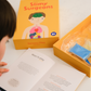 An adult and child are engaged in a science experiment at the kitchen counter, using a beaker and colorful powders. A yellow box labeled "Emotions Part 2: Discovering our Body's Clues Program" by The Play Way sits nearby. Both are focused and interested in the activity.
