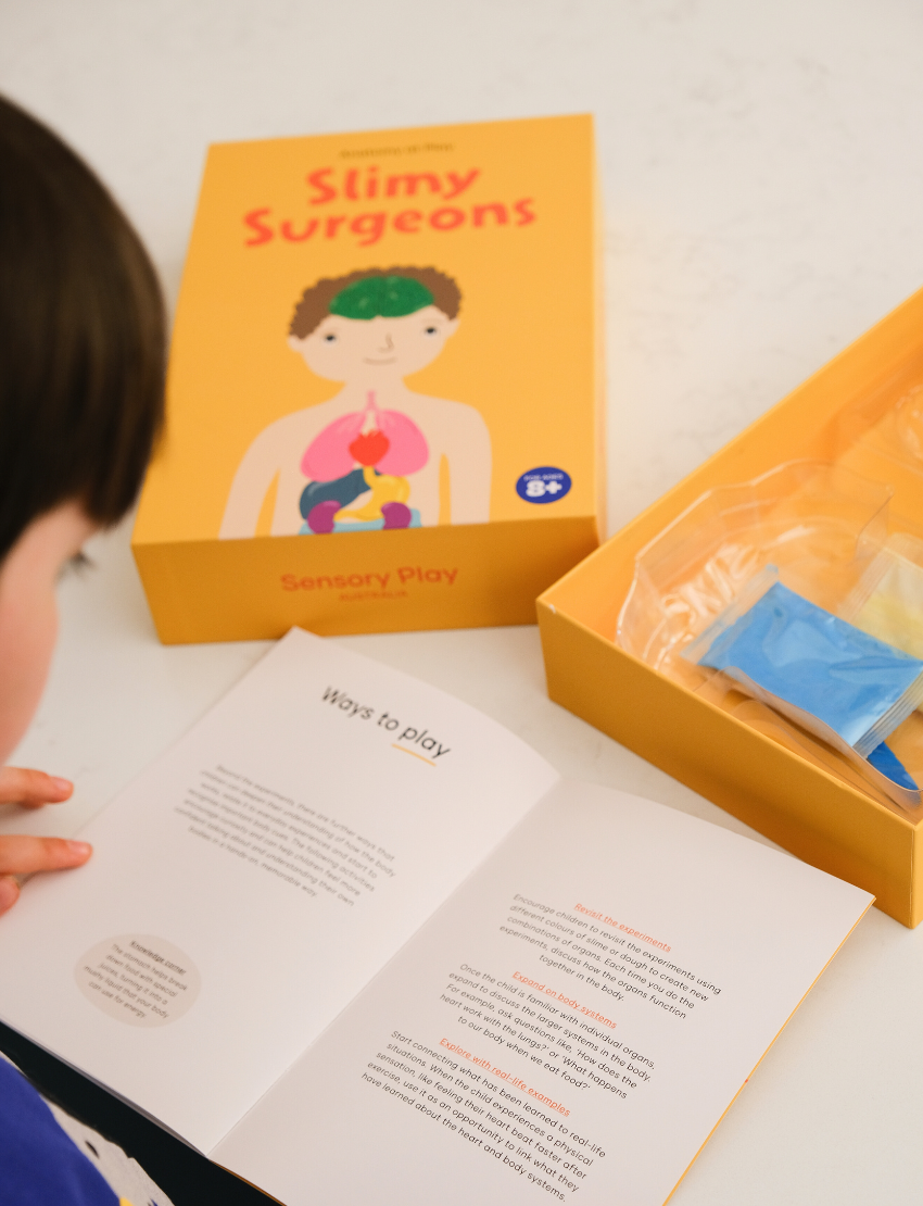 An adult and child are engaged in a science experiment at the kitchen counter, using a beaker and colorful powders. A yellow box labeled "Emotions Part 2: Discovering our Body's Clues Program" by The Play Way sits nearby. Both are focused and interested in the activity.