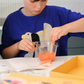 An adult and child are engaged in a science experiment at the kitchen counter, using a beaker and colorful powders. A yellow box labeled "Emotions Part 2: Discovering our Body's Clues Program" by The Play Way sits nearby. Both are focused and interested in the activity.