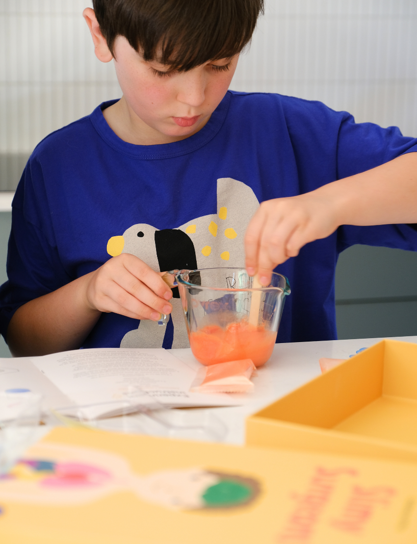 An adult and child are engaged in a science experiment at the kitchen counter, using a beaker and colorful powders. A yellow box labeled "Emotions Part 2: Discovering our Body's Clues Program" by The Play Way sits nearby. Both are focused and interested in the activity.