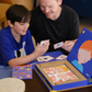 An adult and child are engaged in a science experiment at the kitchen counter, using a beaker and colorful powders. A yellow box labeled "Emotions Part 2: Discovering our Body's Clues Program" by The Play Way sits nearby. Both are focused and interested in the activity.
