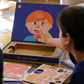 An adult and child are engaged in a science experiment at the kitchen counter, using a beaker and colorful powders. A yellow box labeled "Emotions Part 2: Discovering our Body's Clues Program" by The Play Way sits nearby. Both are focused and interested in the activity.