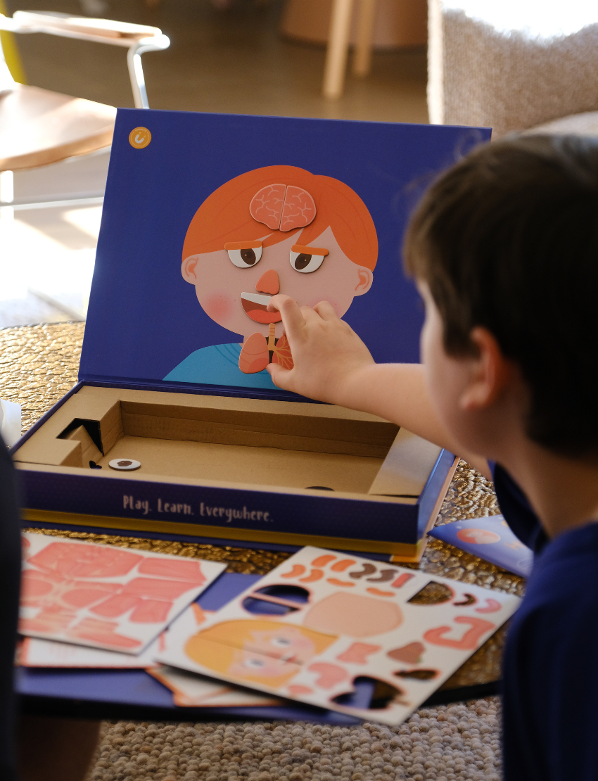 An adult and child are engaged in a science experiment at the kitchen counter, using a beaker and colorful powders. A yellow box labeled "Emotions Part 2: Discovering our Body's Clues Program" by The Play Way sits nearby. Both are focused and interested in the activity.