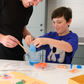 An adult and child are engaged in a science experiment at the kitchen counter, using a beaker and colorful powders. A yellow box labeled "Emotions Part 2: Discovering our Body's Clues Program" by The Play Way sits nearby. Both are focused and interested in the activity.