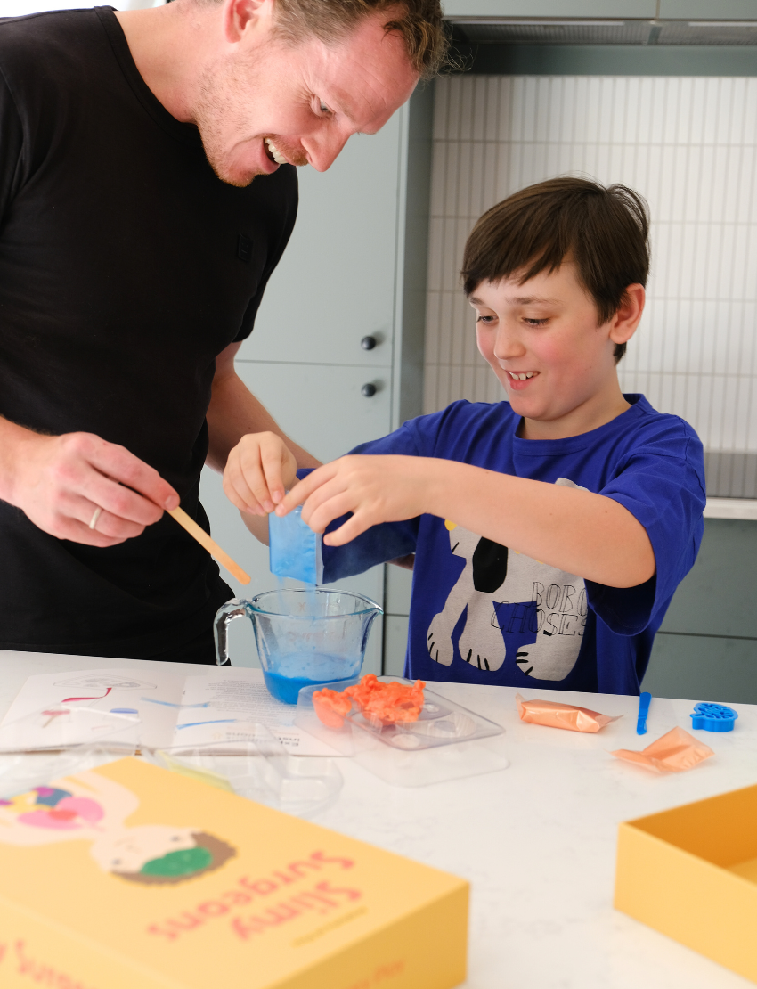 An adult and child are engaged in a science experiment at the kitchen counter, using a beaker and colorful powders. A yellow box labeled "Emotions Part 2: Discovering our Body's Clues Program" by The Play Way sits nearby. Both are focused and interested in the activity.